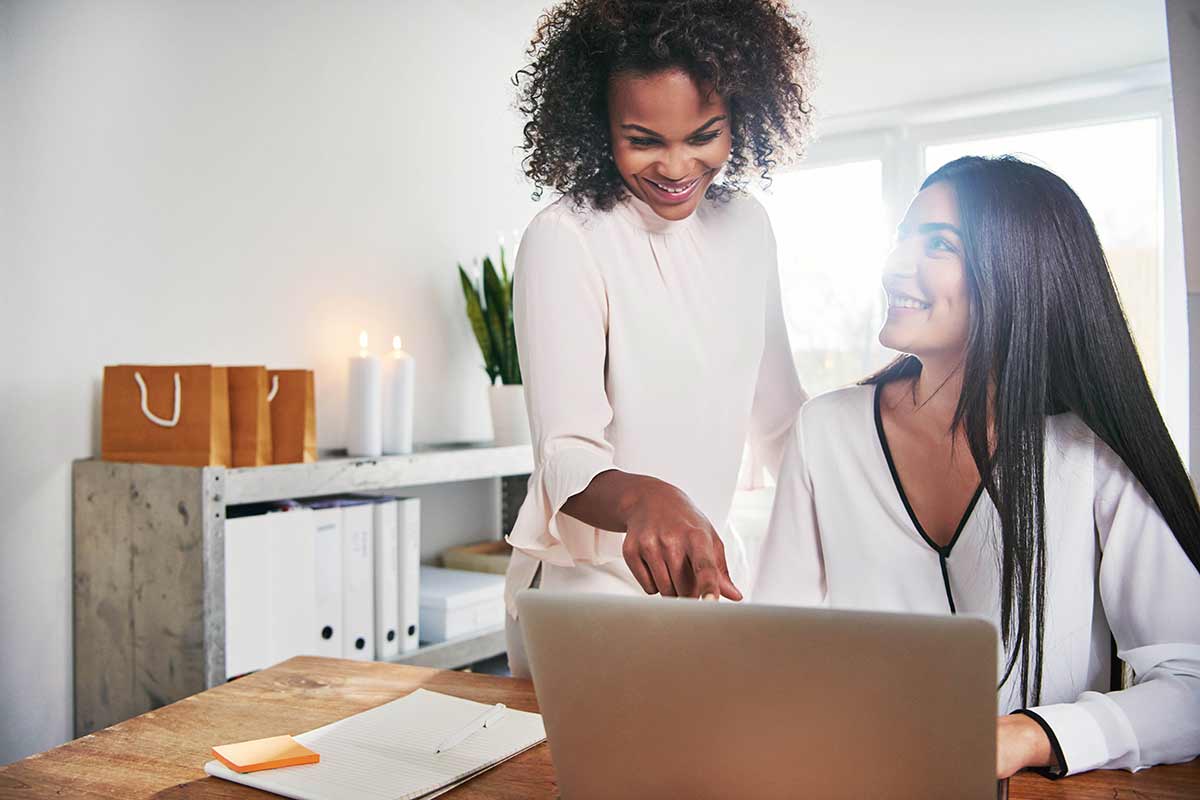 Two women working in a digital marketing agency: one is seated at a laptop, and the other is standing, pointing at the screen. Both appear happy and engaged in their work.